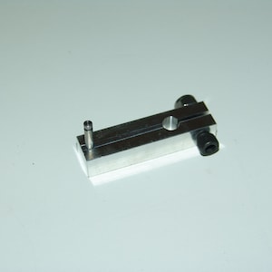 May include: A rectangular silver-colored metal block featuring a central longitudinal slit, a small vertical metal pin at one end, and a black cylindrical screw head with a hexagonal socket integrated into the opposite end of the component.