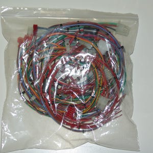 Puede incluir: Un conjunto de cables eléctricos multicolores, incluyendo rojo, verde, azul y amarillo, está enrollado dentro de una bolsa de plástico transparente. Los cables tienen varios conectores y terminales, lo que sugiere que son para un dispositivo o sistema electrónico.