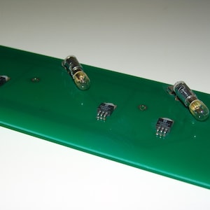 Puede incluir: Una placa de circuito verde con cuatro componentes electrónicos. Dos de los componentes son pequeños circuitos integrados negros. Los otros dos componentes son tubos de vacío de vidrio con carcasas metálicas.