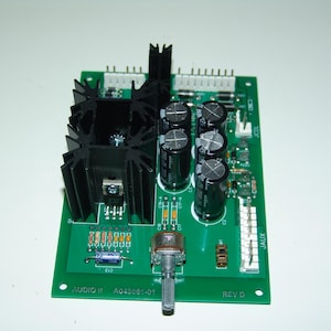 Op de afbeelding: Een groene printplaat met zwarte koellichamen, meerdere condensatoren en een potentiometer. De printplaat is gelabeld met "AUDIO II" en "A04363-01".