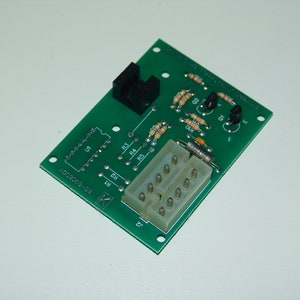 Puede incluir: Una placa de circuito verde con varios componentes electrónicos, incluyendo resistencias, transistores y un conector. La placa tiene varias etiquetas y marcas, como "U1" y "R1-R5". La placa es rectangular con esquinas redondeadas.