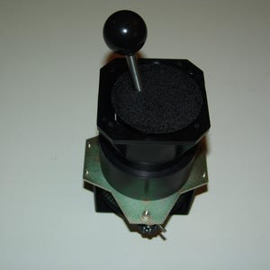 May include: A black joystick with a round, black ball on top. The joystick has a square base with a textured, circular pad. A metal plate is attached to the base. The device is designed for use with electronic equipment.