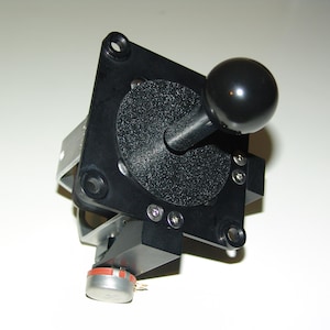 Puede incluir: Un joystick de arcade negro con una perilla redonda negra. El joystick está montado en una base de plástico negro con un potenciómetro plateado.