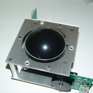 Puede incluir: Un marco de metal plateado con una bola de seguimiento negra en el centro. El marco está unido a una placa de circuito verde con cables y conectores.