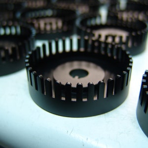 Puede incluir: Primer plano de varios engranajes de plástico negro con un borde exterior dentado y un agujero central. Los engranajes están dispuestos en una fila sobre una superficie azul claro.