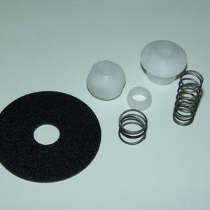 May include: An assortment of mechanical components, including a black circular pad with a central hole, several white plastic pieces, and multiple metal springs. The parts are arranged on a white surface.