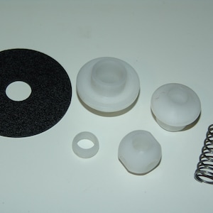 Puede incluir: Un surtido de componentes de plástico blanco y un muelle metálico. Los artículos incluyen una pieza circular negra con un agujero, un anillo y varias formas cilíndricas redondeadas. El muelle es plateado.