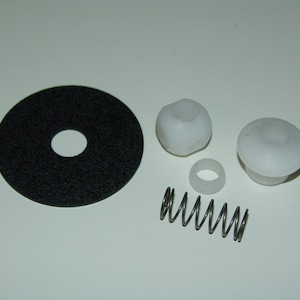 May include: An assortment of small mechanical parts, including a black felt disc with a central hole, two white plastic components, a small white ring, and a silver metal spring. The parts are arranged on a white surface.