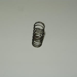 May include: A small, metallic spring with multiple coils. The spring is silver-colored and stands upright against a plain, light-colored background. The spring's design suggests it is a component for machinery or a mechanical device.