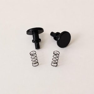 May include: Two black cylindrical fasteners with flat circular heads and two matching metal coil springs. The fasteners feature a stepped design with a wider top section and a narrower base, while the springs are thin, dark, and spiraled in shape.