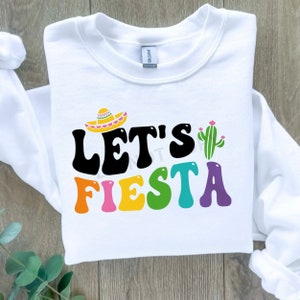 May include: White sweatshirt with the text "Let's Fiesta" in colorful letters. A sombrero and a cactus are above the text.