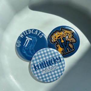 Puede incluir: Tres botones azules y blancos de los Wildcats. Un botón tiene un patrón de cuadros con el texto "Kentucky Wildcats". Otro botón tiene un logotipo de gato montés de dibujos animados. El tercer botón tiene una bota y el texto "Go Wildcats".