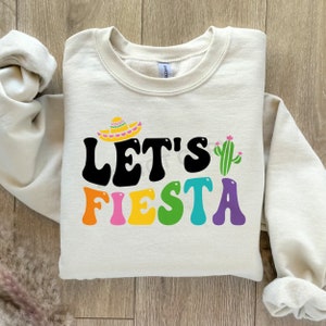 May include: A cream colored sweatshirt with the text "Let's Fiesta" in colorful letters. A sombrero and cactus are above the text.