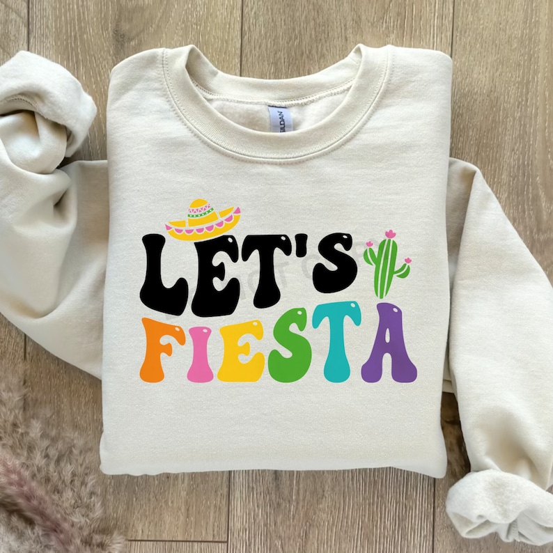 May include: A cream colored sweatshirt with the text "Let's Fiesta" in colorful letters. A sombrero and cactus are above the text.