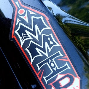 May include: A black and red sticker with white accents. The sticker has a unique shape and features stylized text and symbols. The sticker is on a black surface, possibly a vehicle.