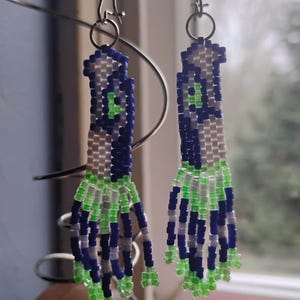 May include: A pair of beaded earrings with a unique design. The earrings feature a combination of blue, white, and green beads. The top portion is primarily blue and white, transitioning to green at the bottom. The earrings hang from silver-colored hooks.