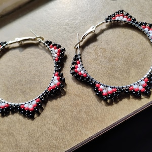 May include: A pair of circular hoop earrings featuring a scalloped edge design. The metal hoops are adorned with small, round beads in a repeating pattern of black, white, and red colors, creating a textured, multi-colored border around the frame.