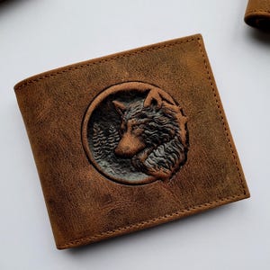 May include: Brown leather wallet with a detailed embossed wolf design within a circular frame. The wallet has a bi-fold design and a textured finish. The wolf is set against a backdrop of trees. The wallet is a rustic accessory.