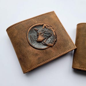 May include: Two brown leather wallets. One wallet features a detailed wolf head design within a circular frame. The wolf is set against a backdrop of trees. The wallets are made of textured leather and have visible stitching.