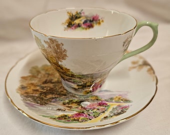 May include: A vintage teacup and saucer set with a delicate floral and landscape design. The cup has a light green handle and a gold rim. The saucer features a similar design with a gold rim.