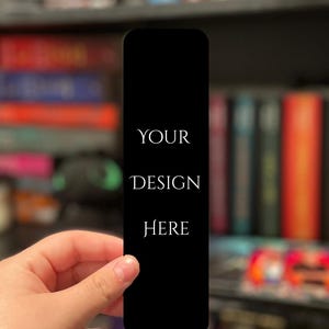 May include: A black bookmark with rounded corners, held by a hand. The text "YOUR DESIGN HERE" is printed in white. The background is a blurred bookshelf with various books.