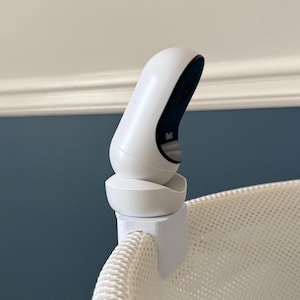 Puede incluir: Un monitor para bebés blanco con un diseño curvo está sujeto a una superficie de malla blanca. El monitor tiene una pantalla oscura y está montado en un clip blanco. El fondo presenta una pared azul y molduras blancas.