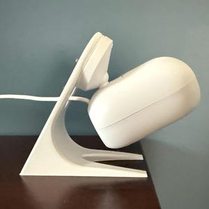 May include: A white, modern-style device with a curved stand and a rounded top. The device is positioned on a dark brown surface against a blue-green wall. A white cord extends from the device.