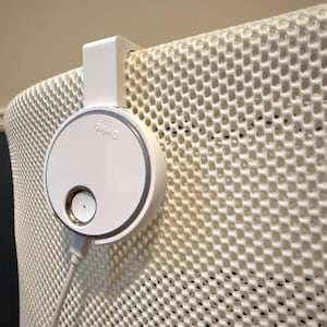 Puede incluir: Un dispositivo blanco y circular con el texto "Omia" está fijado a una superficie blanca con patrón de panal. El dispositivo tiene un pequeño botón y un cable. El dispositivo está montado con un soporte blanco.