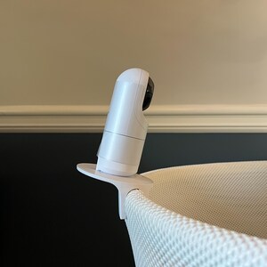 K&ouml;nnte beinhalten: Wei&szlig;er Babymonitor mit einer Kameraobjektiv, das auf einem wei&szlig;en St&auml;nder montiert ist. Der Monitor ist auf einem wei&szlig;en Stubenwagen positioniert.