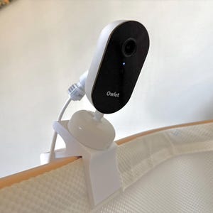 May include: A white and black baby monitor with the brand name "Owlet" printed on the front. The monitor is attached to a white clip, which is secured to the edge of a cot. The monitor has a camera lens and a small blue light.