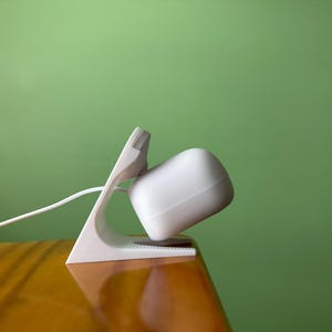 Pode incluir: Uma base de carregamento branca com um dispositivo de formato quadrado e arredondado. A base tem uma base triangular e um braço de suporte curvo. Um cabo branco se estende do dispositivo. O fundo é verde liso.