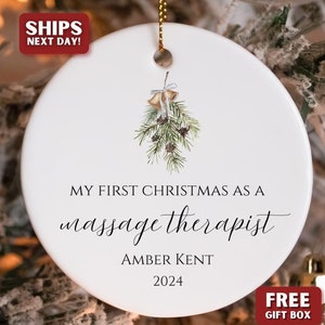 May include: White ceramic Christmas ornament with a green pine sprig and gold bells. The ornament reads "My First Christmas as a massage therapist Amber Kent 2024".