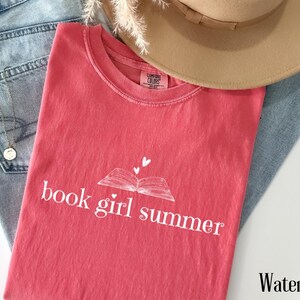 May include: A watermelon-colored t-shirt with the text "book girl summer" and a book graphic. The shirt is laid on top of blue jeans and a tan hat. A gold necklace is visible.