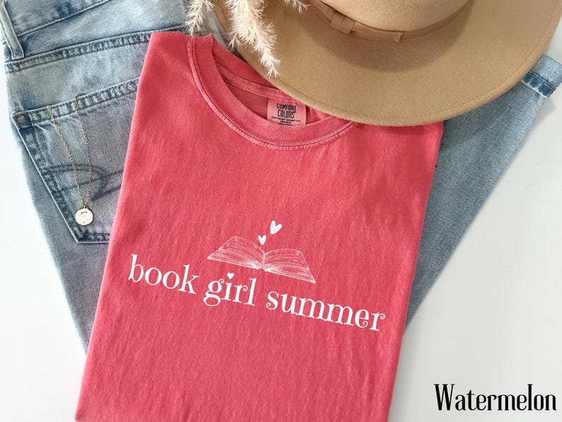 May include: A watermelon-colored t-shirt with the text "book girl summer" and a book graphic. The shirt is laid on top of blue jeans and a tan hat. A gold necklace is visible.