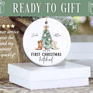 Op de afbeelding: Witte keramische kerstversiering met een boom, cowboylaarzen en de tekst "First Christmas hitched". De namen "Zach" en "Allie" staan boven de laarzen. De versiering ligt op een witte doos.