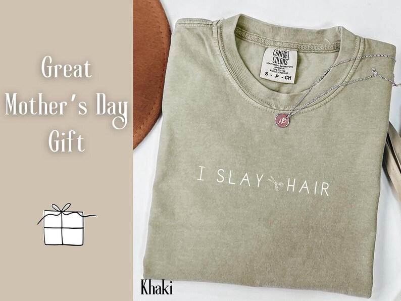 May include: Khaki green t-shirt with the text "I SLAY HAIR" printed on the front. The shirt has a distressed look and a small tag with the text "COMFORT COLORS" on the front.