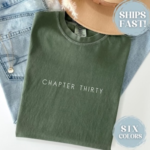 May include: An olive green t-shirt with the text "CHAPTER THIRTY" in white. The shirt is laid on top of light blue jeans, a gold necklace, and a tan hat. The image also includes a graphic that says "SIX COLORS".