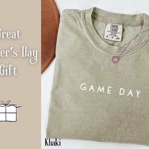 May include: Khaki green t-shirt with the text "GAME DAY" printed in white. The shirt has a crew neck and a tag that says "COMFORT COLORS" with the size "S - P - CH".