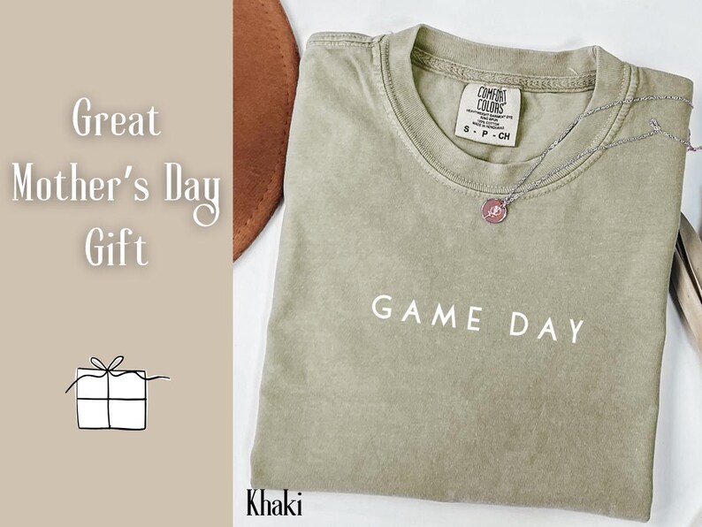 May include: Khaki green t-shirt with the text "GAME DAY" printed in white. The shirt has a crew neck and a tag that says "COMFORT COLORS" with the size "S - P - CH".