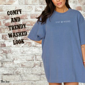 以下が含まれることがあります： 前面に「STAY WEIRD」とプリントされた、ブルーデニムカラーのTシャツ。 シャツはウォッシュ加工で、快適でトレンディと表現されています。 背景はレンガの壁です。
