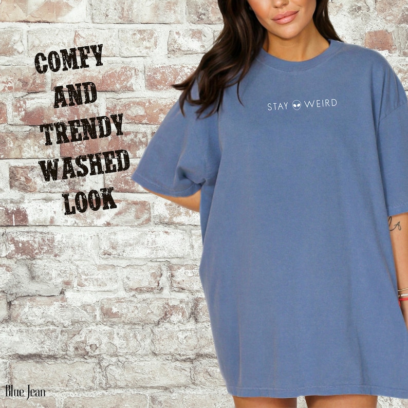 以下が含まれることがあります： 前面に「STAY WEIRD」とプリントされた、ブルーデニムカラーのTシャツ。 シャツはウォッシュ加工で、快適でトレンディと表現されています。 背景はレンガの壁です。