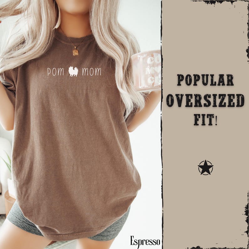 May include: A brown, oversized t-shirt with the words "pom mom" and a Pomeranian dog graphic in white. The shirt has a relaxed fit and is made of soft material. The text "POPULAR OVERSIZED FIT!" is on the right side of the image.