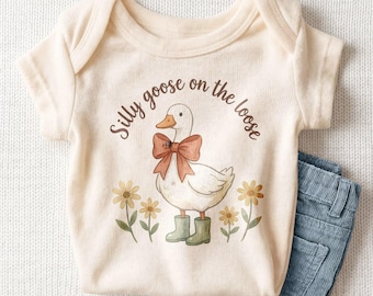 Puede incluir: Un body de bebé de manga corta color crema con el gráfico de una oca blanca que lleva un lazo naranja grande y botas de agua verdes, rodeada de flores amarillas. El texto "Silly goose on the loose" está impreso en marrón sobre la oca.