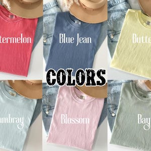 May include: A collection of six t-shirts in various colors: Watermelon, Blue Jean, Butter, Chambray, Blossom, and Bay. Each shirt is folded and displayed with a hat and denim jeans. The word "COLORS" is in the center.