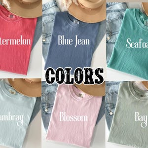 K&ouml;nnte beinhalten: Sechs einfarbige T-Shirts in verschiedenen Farbt&ouml;nen wie Wassermelone, Blue Jean, Seafoam, Chambray, Blossom und Bay. Jedes Shirt ist gefaltet und wird mit einem Hut und Jeans pr&auml;sentiert. Das Wort "COLORS" ist in der Mitte zu sehen.