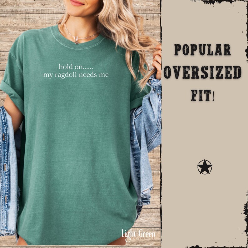 May include: Light green oversized t-shirt with the text "hold on..... my ragdoll needs me" in white. The shirt is a relaxed fit and has rolled-up sleeves. The words "Popular Oversized Fit!" are printed on the right side of the image.
