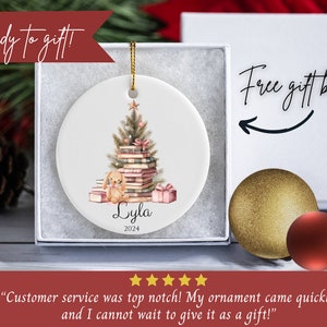 May include: A personalized Christmas ornament featuring a watercolor illustration of a stack of books with a Christmas tree on top. A small bunny sits on the books. The ornament is white with the name "Lyla" and the year "2024" printed on it. The ornament comes in a white gift box with the text "Free gift box!" printed on it.