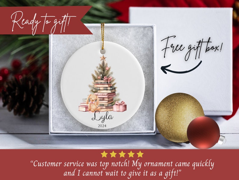 May include: A personalized Christmas ornament featuring a watercolor illustration of a stack of books with a Christmas tree on top. A small bunny sits on the books. The ornament is white with the name "Lyla" and the year "2024" printed on it. The ornament comes in a white gift box with the text "Free gift box!" printed on it.