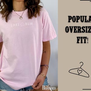 K&ouml;nnte beinhalten: Hellrosa Oversized-T-Shirt mit wei&szlig;em Schriftzug "LAKE BUM". Das Shirt hat kurze &Auml;rmel und eine lockere Passform. Das Bild enth&auml;lt auch den Text "POPULAR OVERSIZED FIT!" und eine Kleiderb&uuml;gel-Grafik.