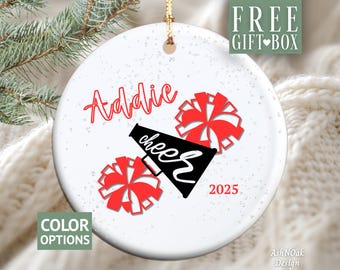 May include: White ceramic ornament with red text that says "Addie" and "cheer". Two red pom-poms and a black megaphone are also featured. The year "2025" is in red. The ornament has a gold string for hanging.